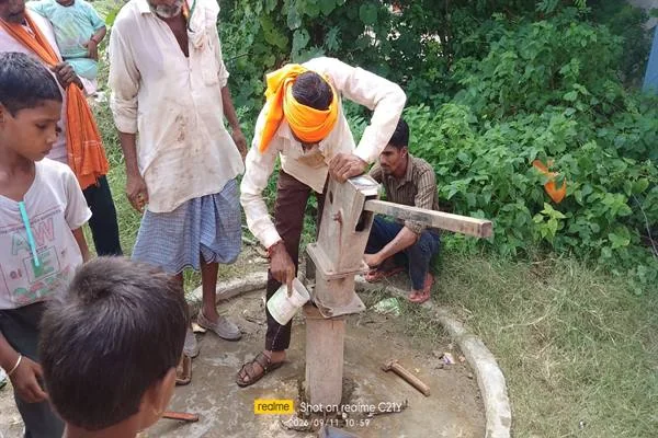 हैंडपंप के पास खड़ा व्यक्ति एक सफेद डिब्बे से सामग्री डालते हुए, आसपास ग्रामीण मौजूद