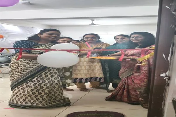 छात्रावास के एक कक्ष के दरवाजे पर साड़ी और सलवार-सूट पहने महिलाएं व छात्राएं गुलाबी रिबन काटती हुईं, दरवाजे पर सफेद-गुलाबी गुब्बारे और तोरण लगे हैं।